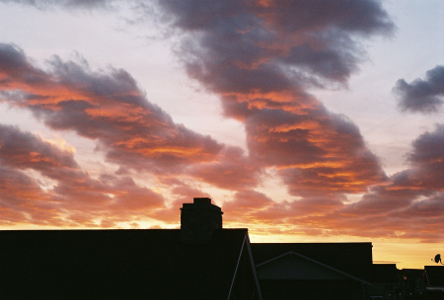 [The outlines of the tops of the buildings are dark. The sky at the horizon is shades of yellow. The rest of the sky is white-blue except for the clouds which are mostly dark grey except for segments of orange on parts of the lower levels.]